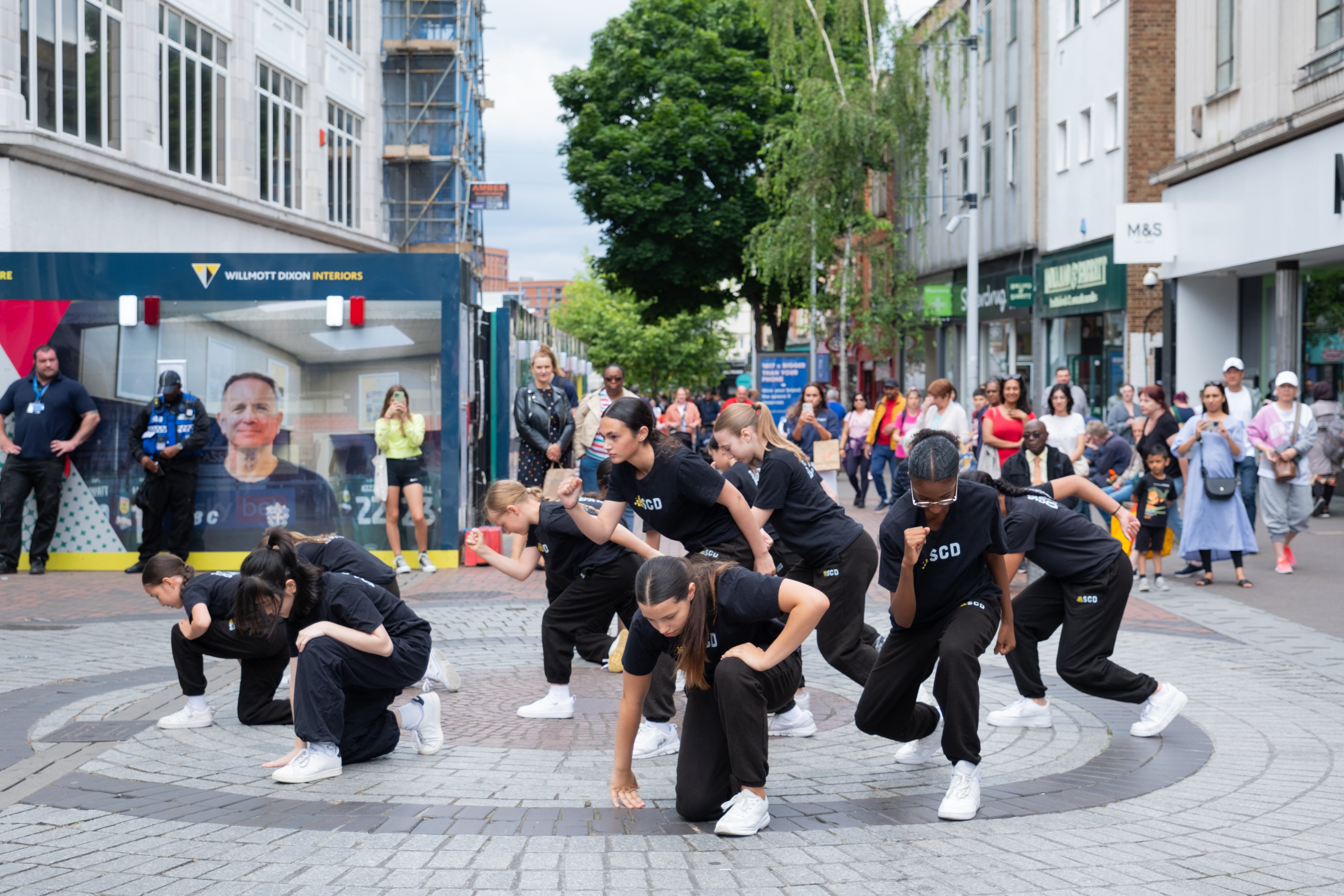 Sutton Community Dance (SCD) group performing in Sutton High Street, outside Marks & Spencer shop, with members of public watching on.