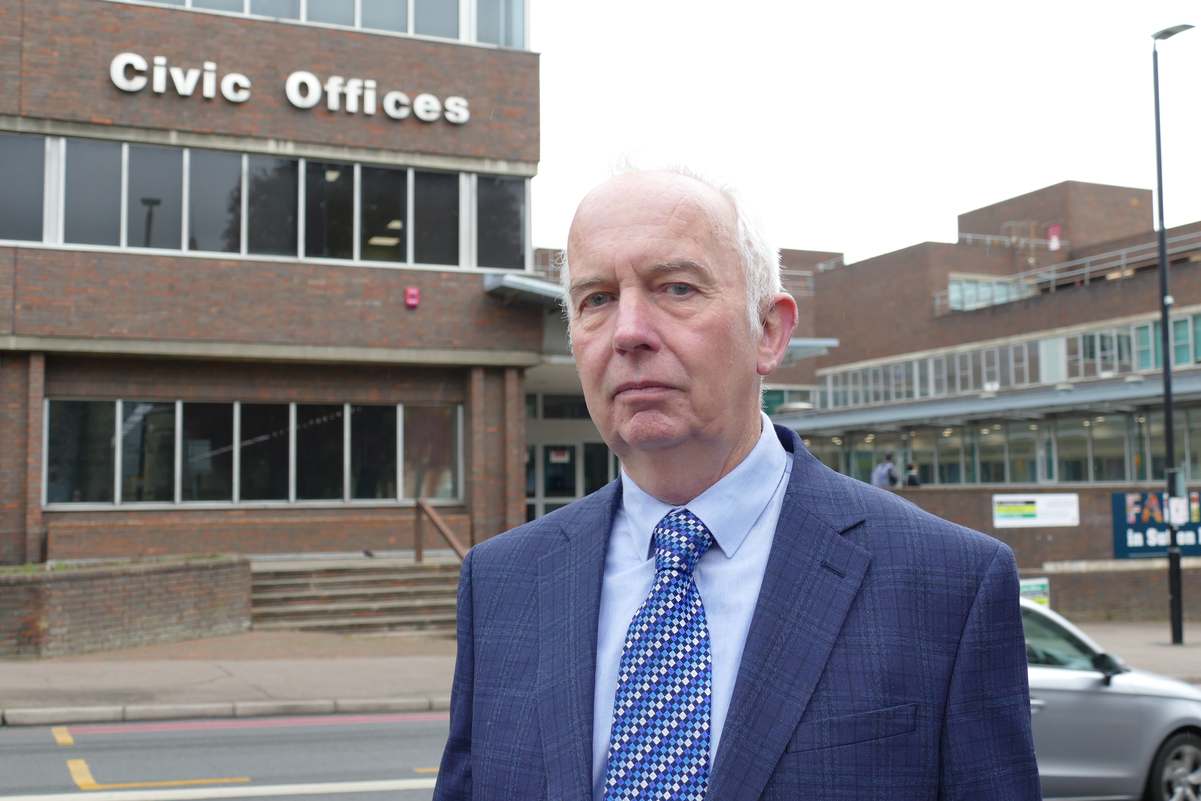 Council Leader Barry Lewis standing in front of Sutton Civic Offices wearing a blue suit jacket and blue patterned tie.