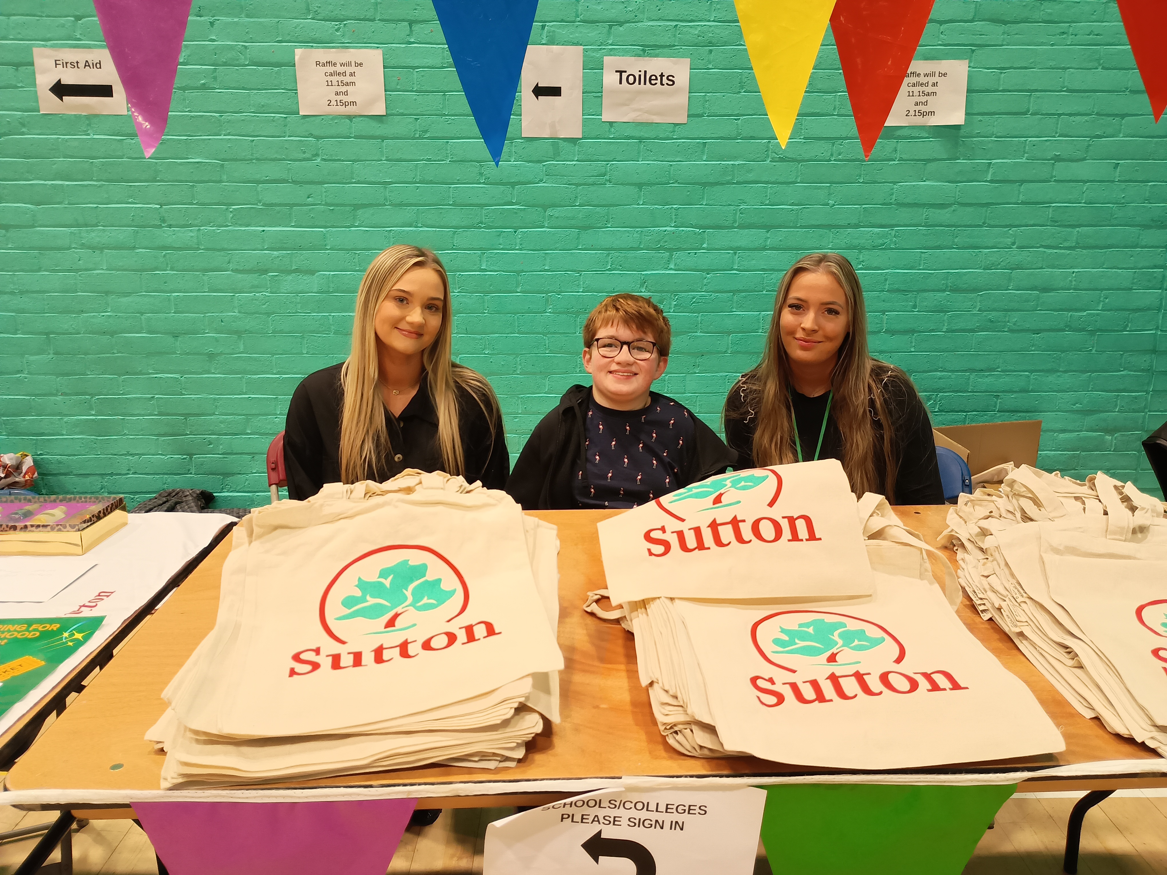 image of a young volunteer and 2 women sat at a sign in table containing lots of tote bags featuring the sutton logo