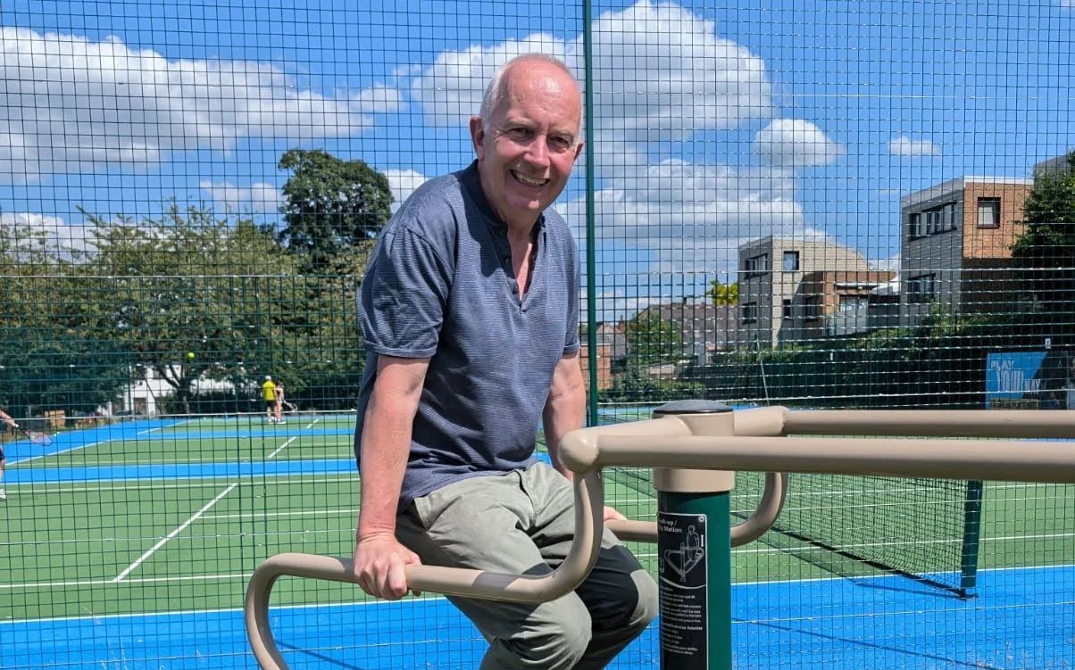 Leader of the Council, Councillor Barry Lewis using outdoor gym equipment with tennis courts behind him
